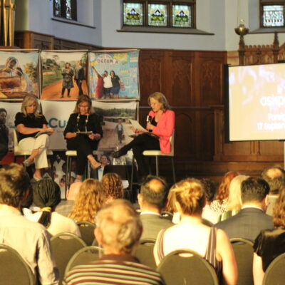 Un panel de trois intervenants présente sur une petite scène devant un public dans une salle aux murs en bois.