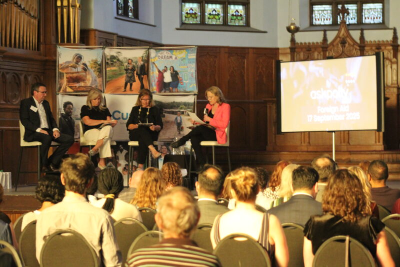 A panel of three speakers presents on a small stage in front of an audience in a wood paneled room.