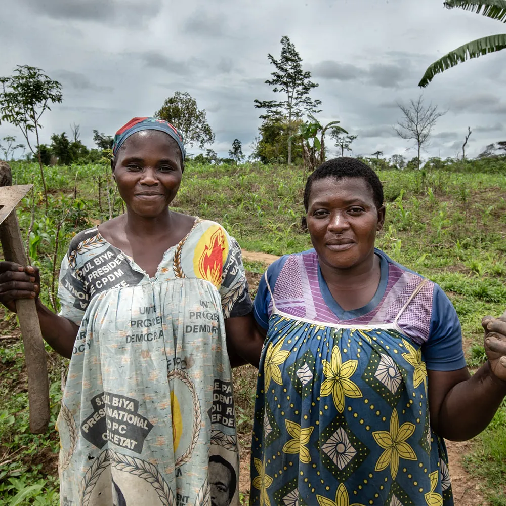 Two women standing together outdoors, smiling, with farmland behind them.