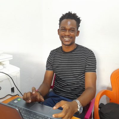 A young African man in a striped t-shirt sitting at a computer and smiling at the camera