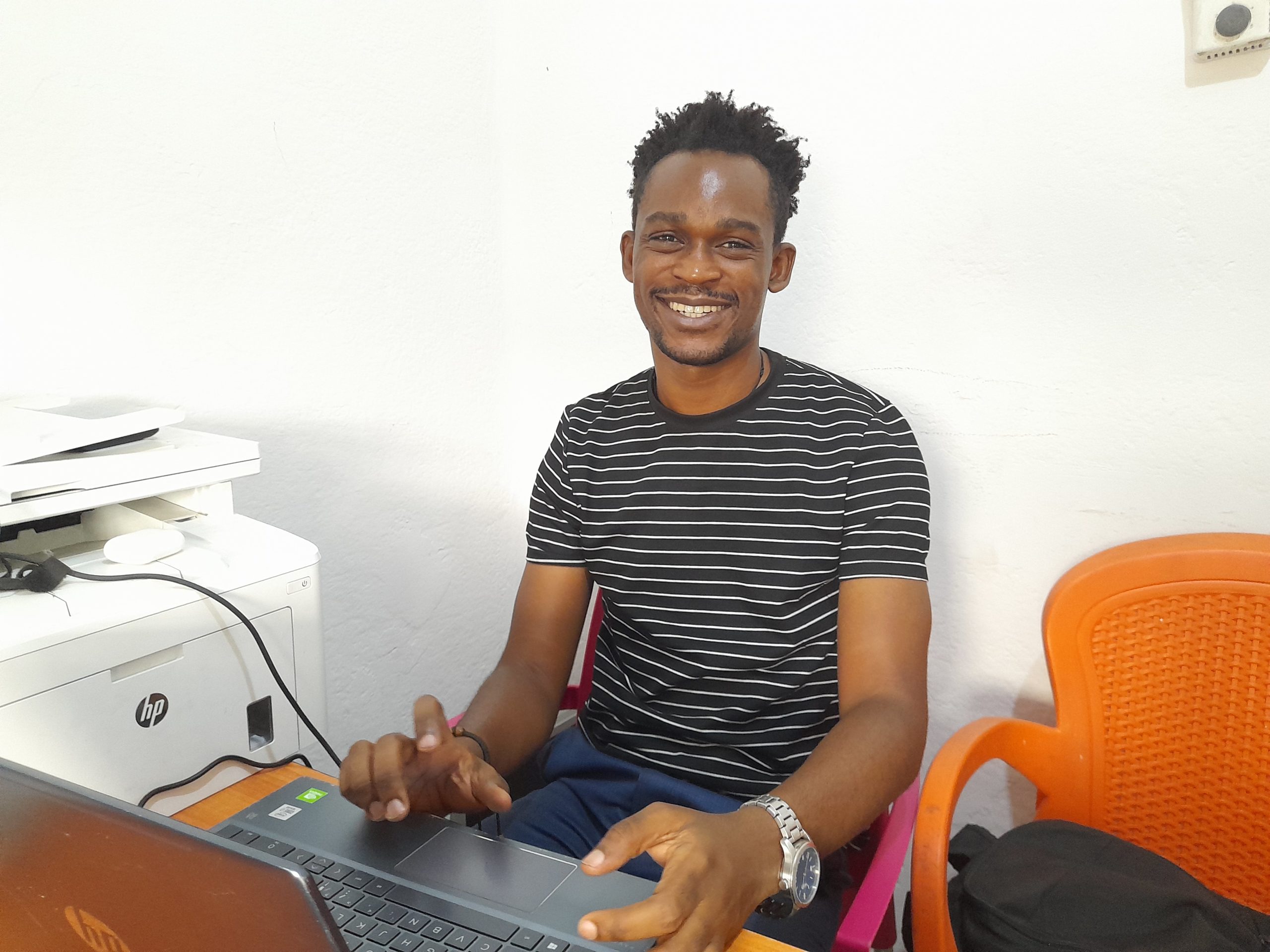 A young Black man in a striped t-shirt sitting at a computer and smiling at the camera