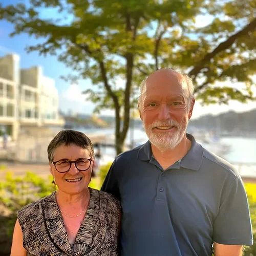 “Smiling older couple standing outdoors in front of trees and water, with buildings in the background.