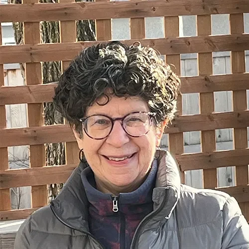 Older woman with curly hair and glasses smiling in front of a wooden fence.