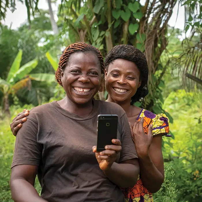 Two women in Cameroon, smiling and taking a selfie, capture the lush green backdrop on their smartphones. Cuso International.