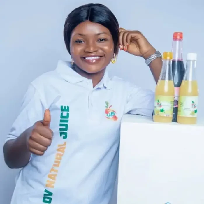 A woman in a white shirt smiles, giving a thumbs up next to Cuso International juice bottles in Cameroon.