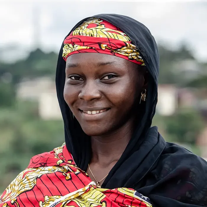 A smiling person in a colorful headscarf and patterned clothing, with the blurred outdoors of Cameroon behind them.