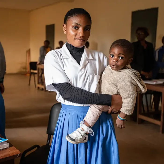 A woman in a white coat holds a child, standing in a Cuso International classroom in Cameroon with desks and people.