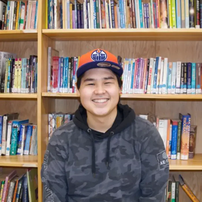 A person in a cap smiles while sitting in front of bookshelves and colorful books, dreaming of the Northwest Territories.