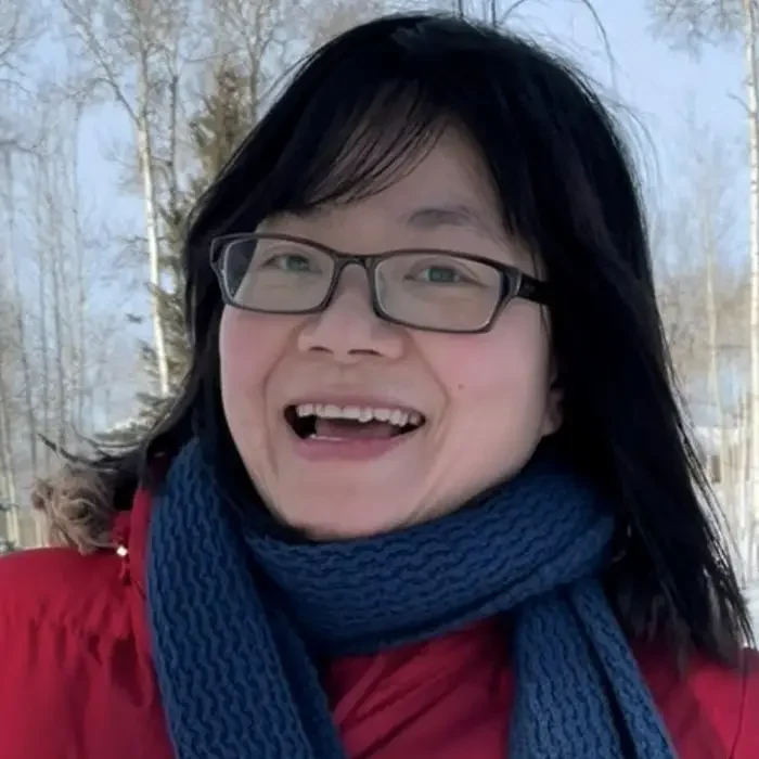 A smiling person with glasses and a blue scarf stands outdoors in the Northwest Territories beneath a clear sky.