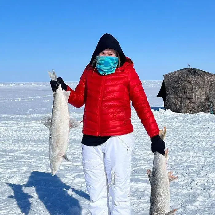 Person in red jacket holds two large fish on a snowy Northwest Territories landscape, with a tent in the background.