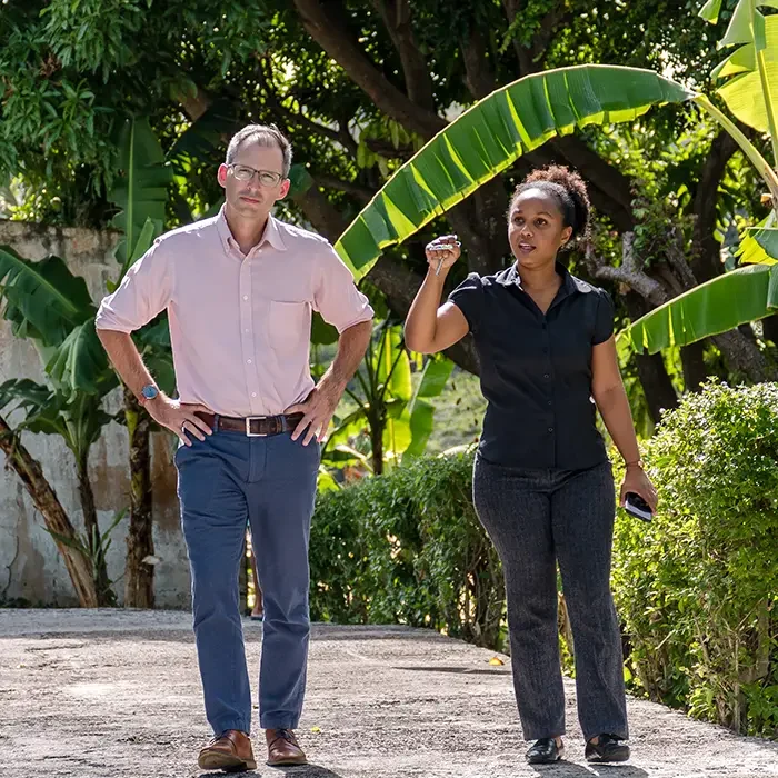 Two people walking outdoors, one gesturing while talking, amid lush greenery in the Caribbean.