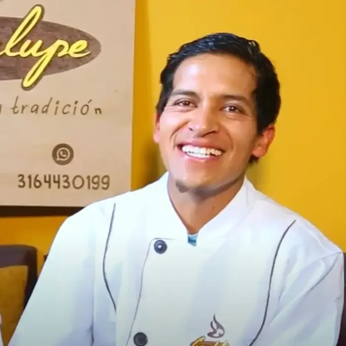 Smiling person in a white chef's coat, sitting indoors with a yellow backdrop and Cuso International signage visible.