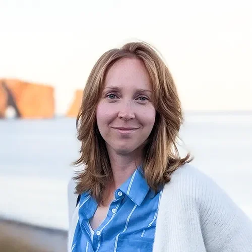 Woman with light brown hair wearing a blue shirt and white sweater, smiling outdoors.