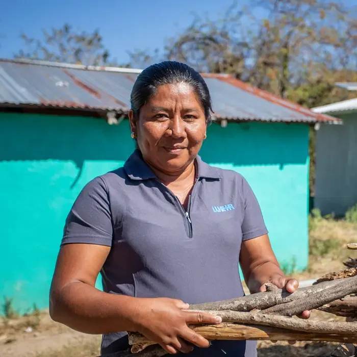 A person in a gray shirt holds firewood in front of a green Cuso International Honduras building with a tin roof.