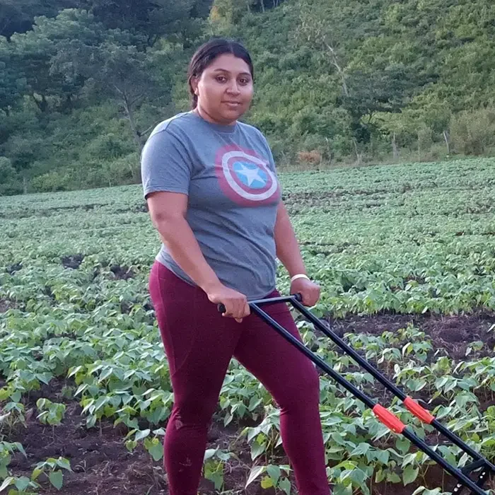 A person in a superhero shirt and maroon pants uses a manual tiller in a lush field, embracing Cuso International's spirit.