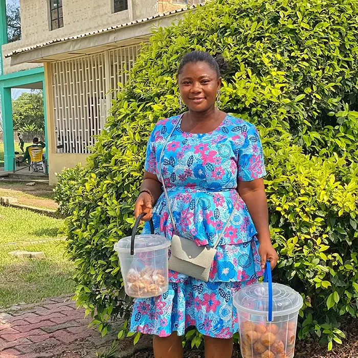 Woman in a floral dress holds two buckets, standing by a leafy bush near Cuso International Nigeria's building.