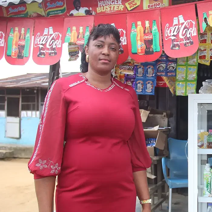 A woman in a red dress stands before a small shop with Coca-Cola banners, showcasing Cuso International Nigeria's impact.