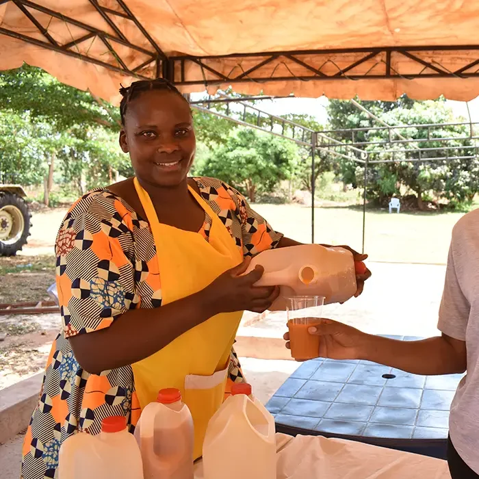 A woman in a colorful dress pours juice into a cup under a canopy, supporting Cuso International's work in Tanzania.