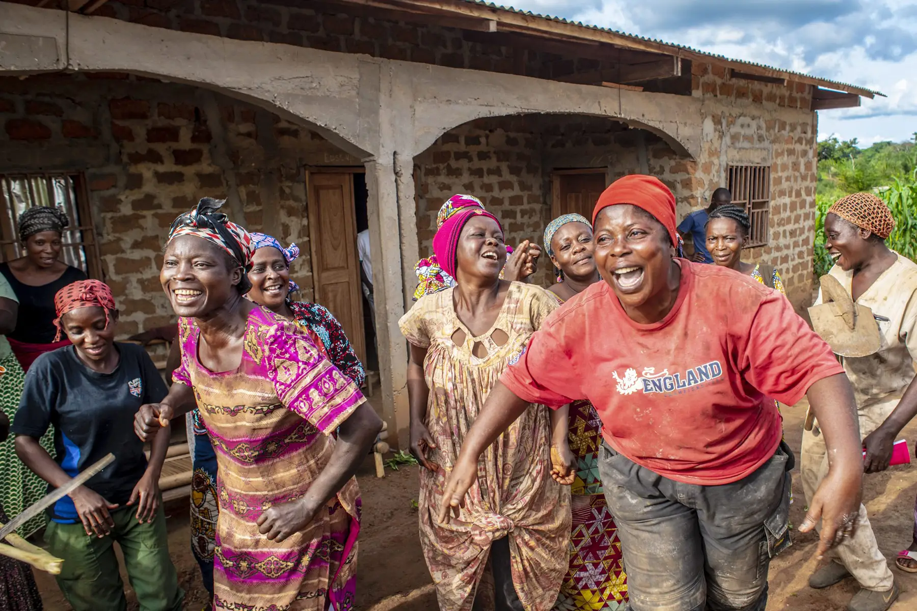 A group of women joyfully dancing and laughing outside a brick building in rural Cameroon.