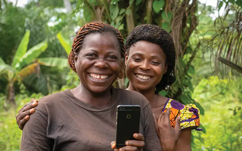 Two people smile at a smartphone in lush greenery, capturing the joy of Cuso International Cameroon.