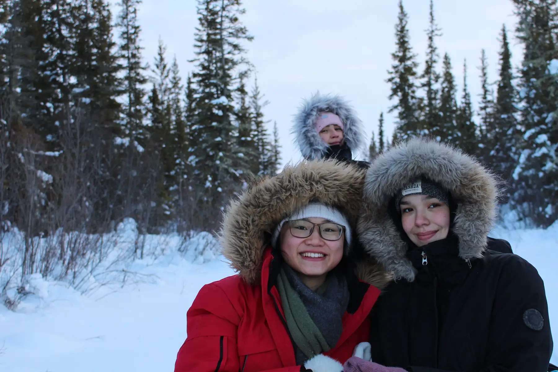 Three people in fur-hooded winter coats smile in a snowy Northwest Territories forest, tall trees towering behind.