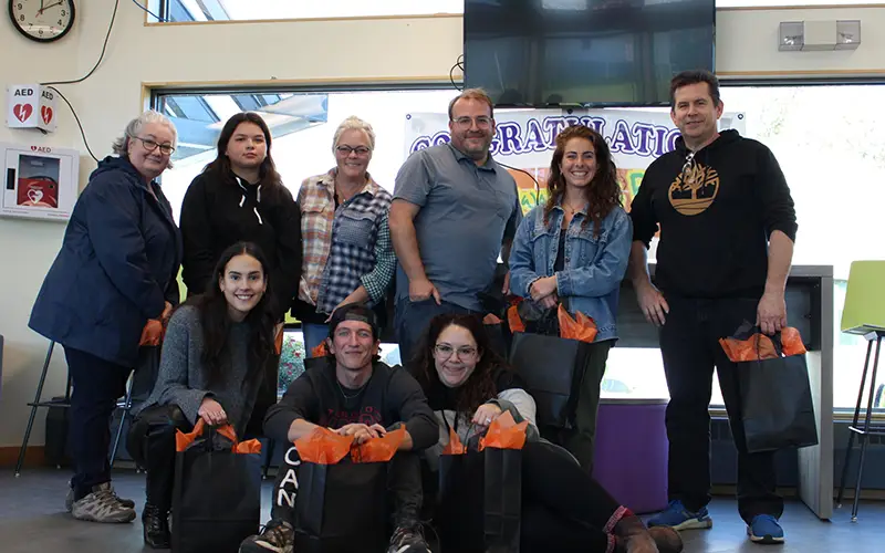 A group of eight people smiling indoors, holding gift bags in the Northwest Territories, with a congratulatory banner.