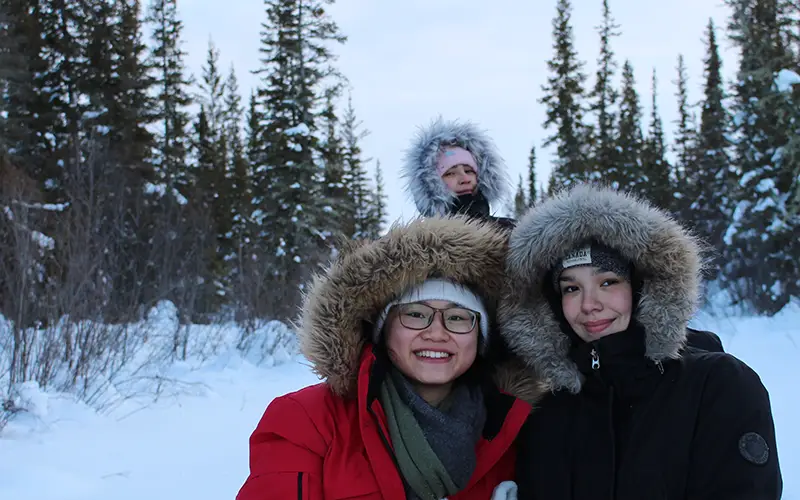 Three people in winter coats and fur-lined hoods smile in a snowy forest setting in the Northwest Territories.