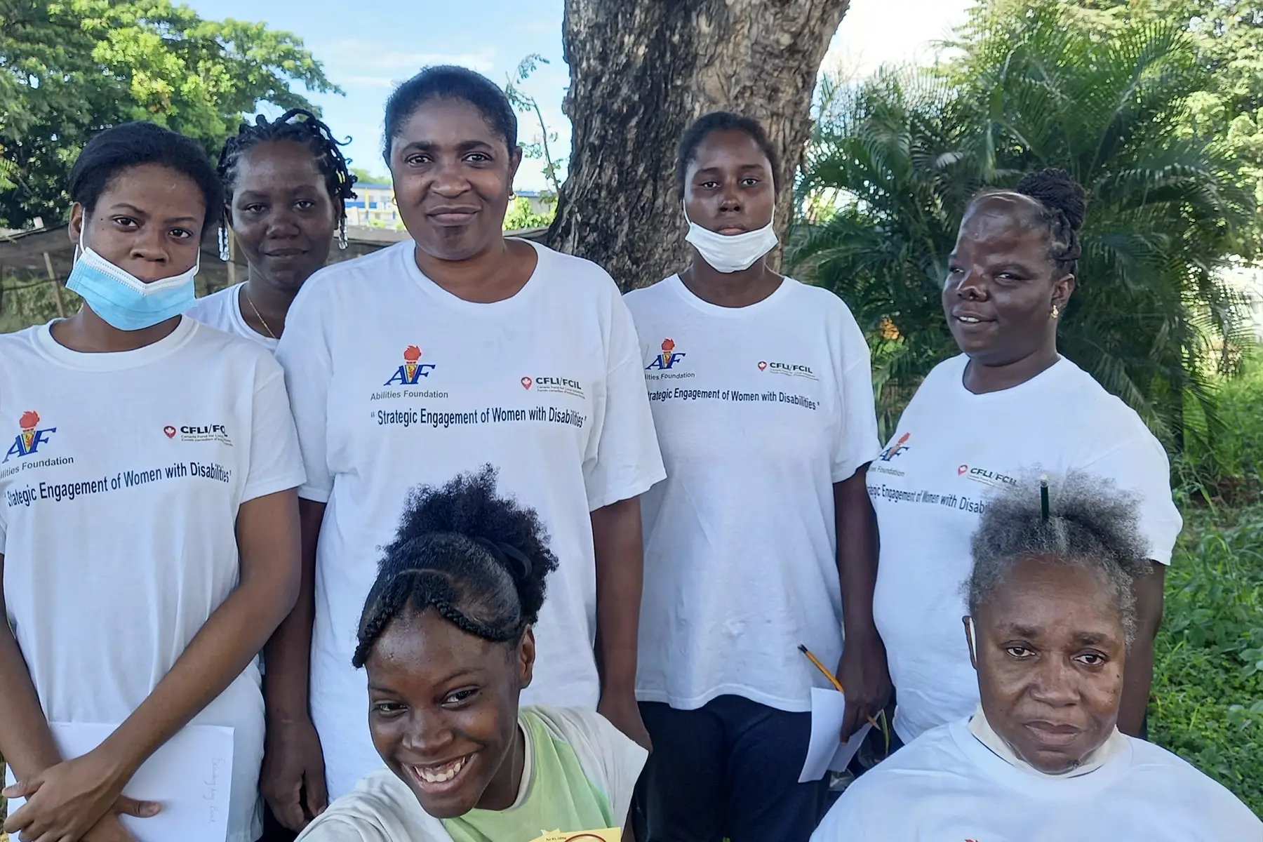 A group of seven women in white shirts smiles outdoors under a large tree, some holding papers, in the Caribbean.