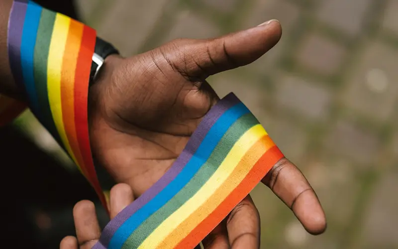 A hand holds a rainbow ribbon, symbolizing LGBTQ+ pride, against a blurred brick pavement in the Caribbean.