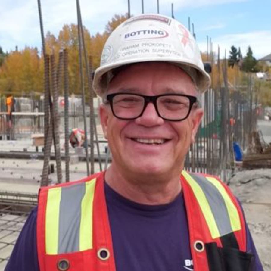 An older man smiling with glasses and a hard hat on a construction site