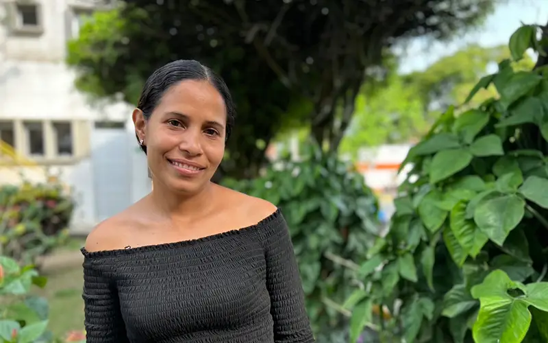 Smiling woman in a black off-shoulder top stands outside, embraced by green foliage in Colombia.