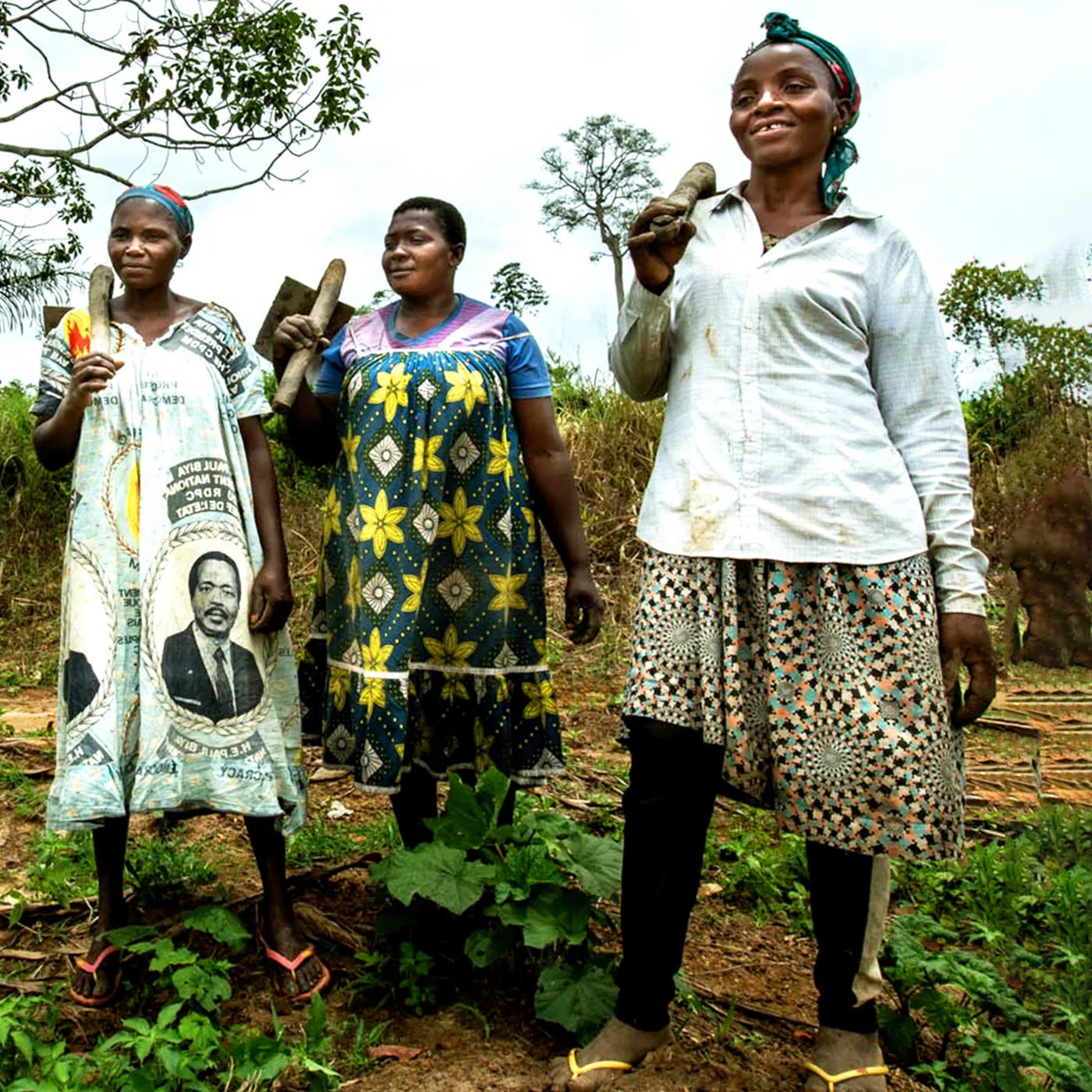 Three Women stand together outdoors in a lush rural landscape, each holding a farming tool over their shoulder. They wear brightly patterned dresses and headscarves, and they smile confidently toward the camera. Tall trees and dense vegetation fill the background on an overcast day.