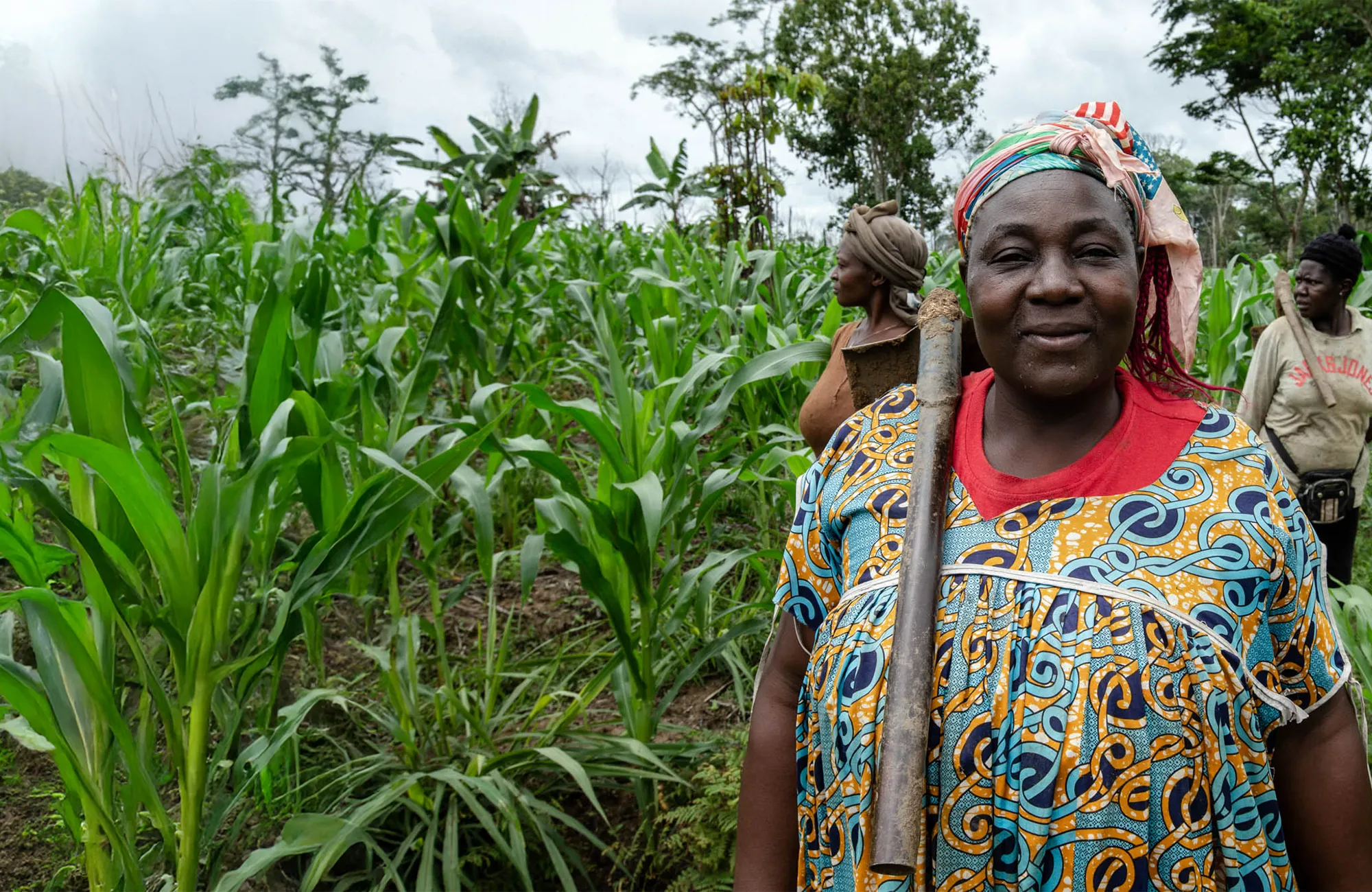 Woman in colourful patterned clothing holding a tool over her shoulder, standing beside tall green crops in a field.