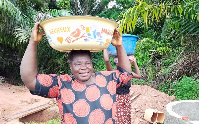 In a lush green setting, a smiling woman carries a large bowl on her head, alongside another with a blue container in the DRC.