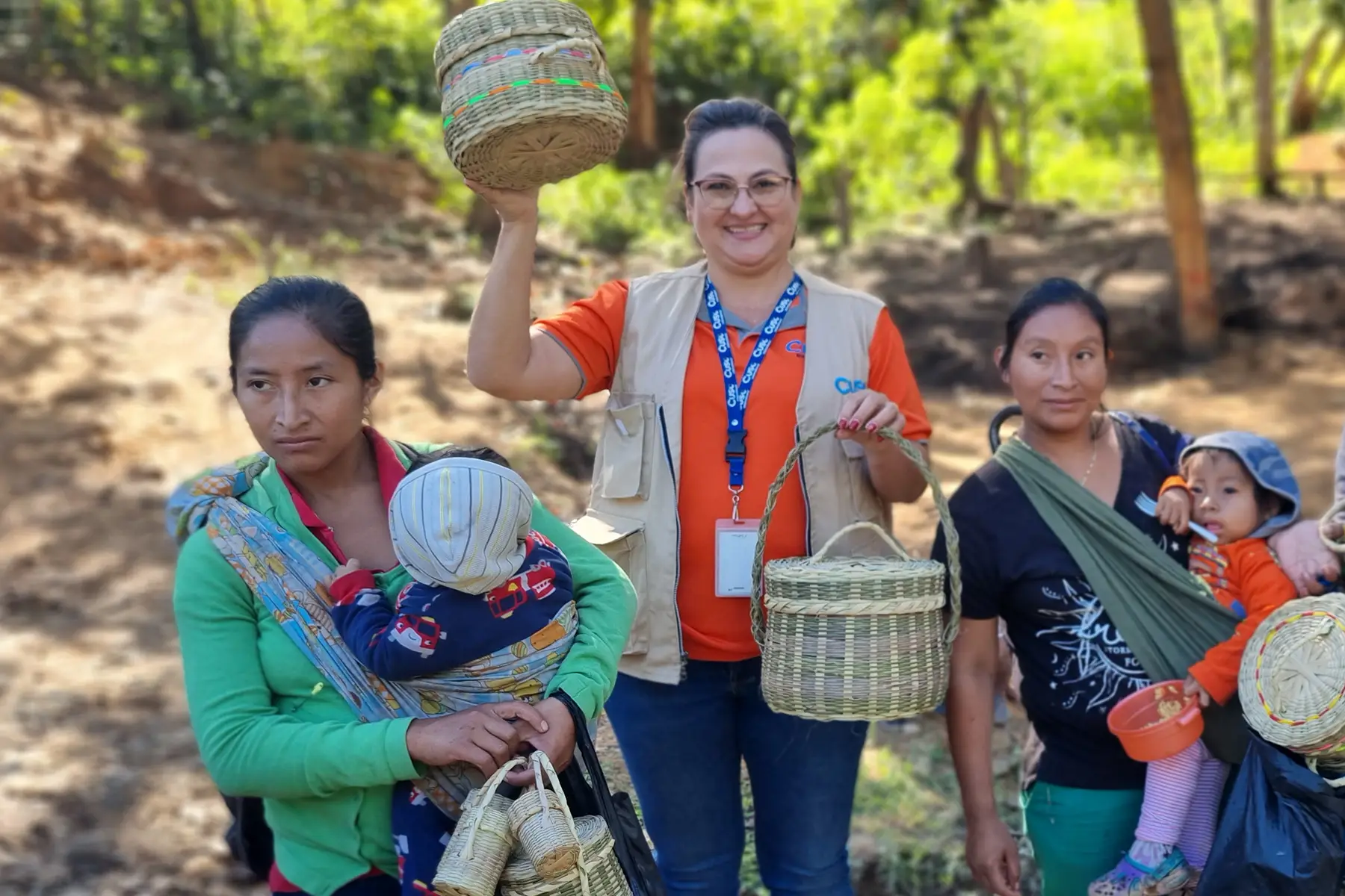 Three women outdoors in Honduras, two with babies in wraps, one lifting baskets, trees behind them. Cuso International.