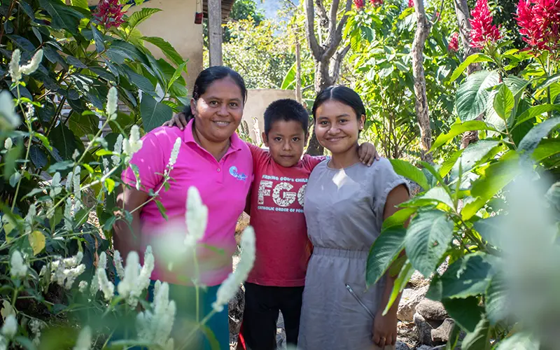 Three people smiling in a lush garden, surrounded by green foliage and vibrant blooms, celebrating Cuso International.