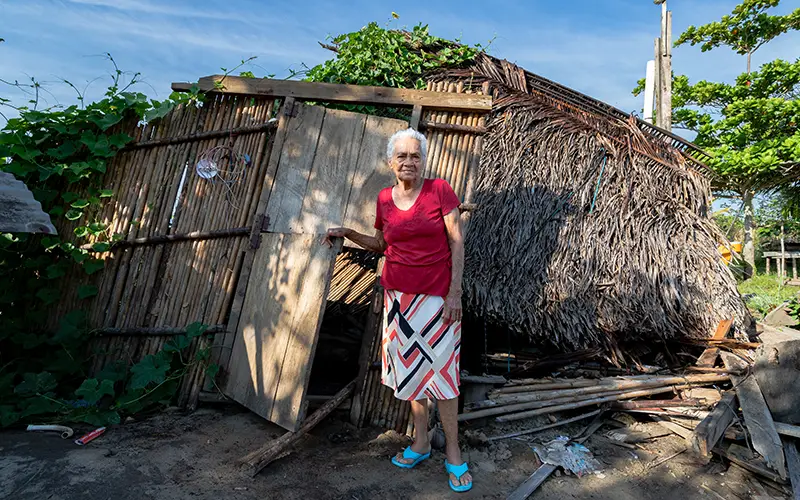 Elderly woman in red shirt, standing by a rustic hut with a thatched roof in Honduras, under a clear blue sky.