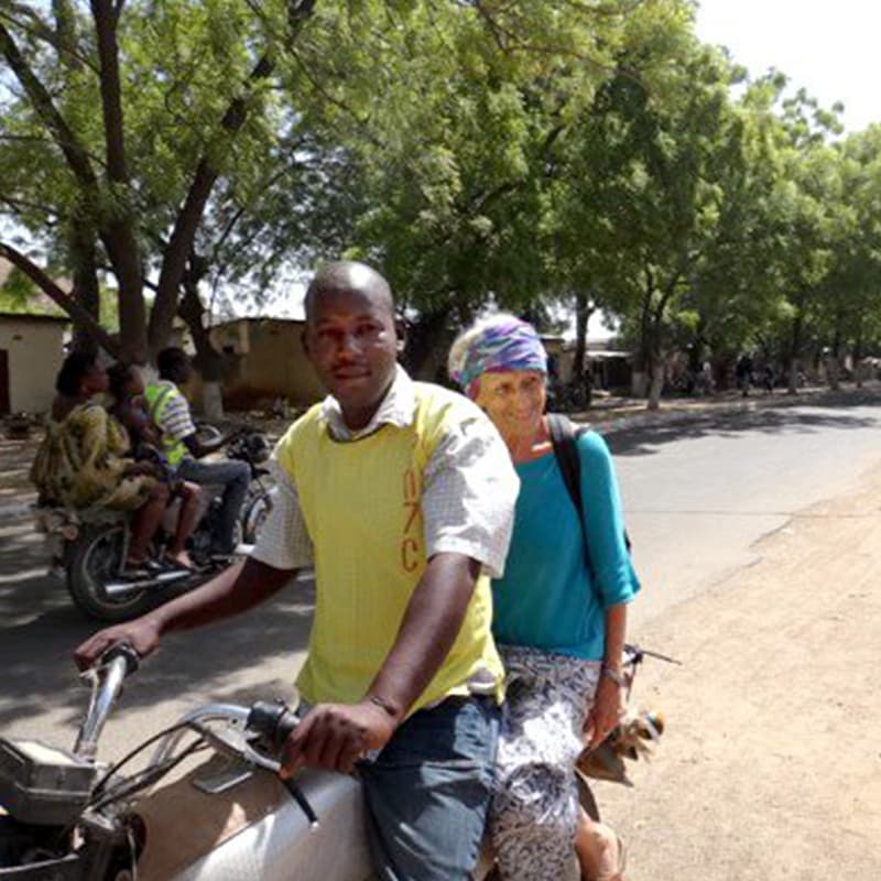 A woman with a turquoise shirt sitting on the back of a motorcycle on a dirt road