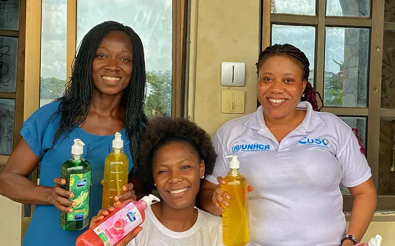 Three women smiling with Cuso International Nigeria bottles of liquid soap outside a building.