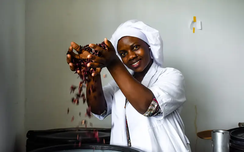 A woman in a white coat and hat smiles, holding dried beans over a container in Tanzania with Cuso International.